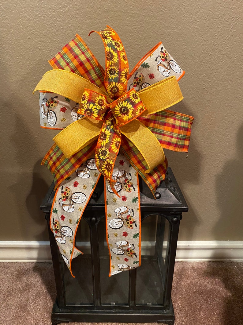 May include: A large, multi-colored bow with a plaid ribbon, a sunflower ribbon, and a ribbon with a bicycle and sunflower pattern. The bow is tied around a black lantern.