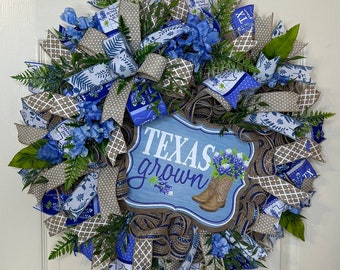 Peut inclure: Une couronne bleue et brune avec une pancarte qui dit "Texas grown" et représente une paire de bottes de cowboy. La couronne est décorée de ruban bleu et blanc, de fleurs bleues et de feuilles vertes.
