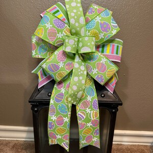 Puede incluir: Un lazo de Pascua verde, blanco y rosa con un patrón de lunares y un diseño de huevos de Pascua coloridos. El lazo está atado alrededor de una linterna negra.