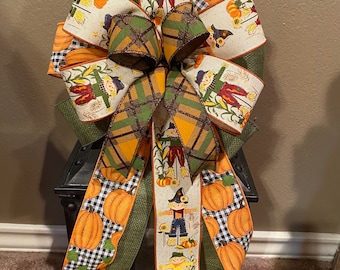 May include: A large, multi-coloured bow with an autumn theme. The bow is made of several different ribbons, including a plaid ribbon, a ribbon with a pumpkin pattern, and a ribbon with a scarecrow pattern. The bow is tied with a green ribbon and is accented with a small, brown bow.