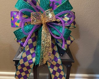 May include: A purple, gold, and green Mardi Gras bow with a checkered pattern. The bow is tied with a gold mesh ribbon and is sitting on top of a black lantern.