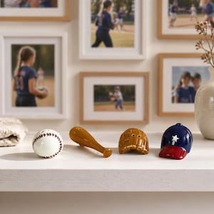 Puede incluir: Un conjunto decorativo con tem&aacute;tica de b&eacute;isbol que incluye una pelota blanca con costuras rojas, un bate marr&oacute;n, un casco marr&oacute;n y una gorra de b&eacute;isbol azul y roja. El conjunto se exhibe sobre una superficie blanca, con fotos enmarcadas de jugadores de b&eacute;isbol en el fondo.