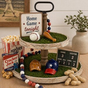 Puede incluir: Una bandeja de madera de dos niveles con decoraciones tem&aacute;ticas de b&eacute;isbol. El nivel superior presenta una pelota de b&eacute;isbol, un bate y un letrero que dice "Home Game tonight". El nivel inferior tiene un guante de b&eacute;isbol, una gorra y un marcador. Tambi&eacute;n hay palomitas de ma&iacute;z, Cracker Jack y cacahuetes.