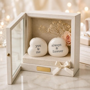 May include: A white wooden display box with a glass door containing two white heart-shaped ceramic stones. The left stone is printed with the text you & me, and the right stone is printed with always & forever. A cream ribbon and dried flowers are included.