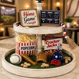 Puede incluir: Una bandeja blanca envejecida de dos niveles con decoraciones tem&aacute;ticas de b&eacute;isbol. Incluye una pelota de b&eacute;isbol, un bate, un guante y una gorra. El nivel superior tiene un letrero que dice "Home Game tonight" y un marcador. Tambi&eacute;n hay una bolsa de palomitas de ma&iacute;z y Cracker Jacks.