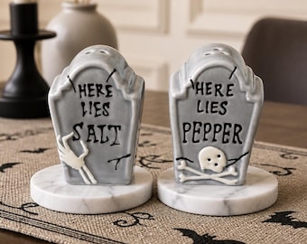 May include: A pair of gray ceramic tombstone-shaped salt and pepper shakers. The salt shaker features a white skeletal hand and the text "HERE LIES SALT". The pepper shaker features a white skull and crossbones and the text "HERE LIES PEPPER".