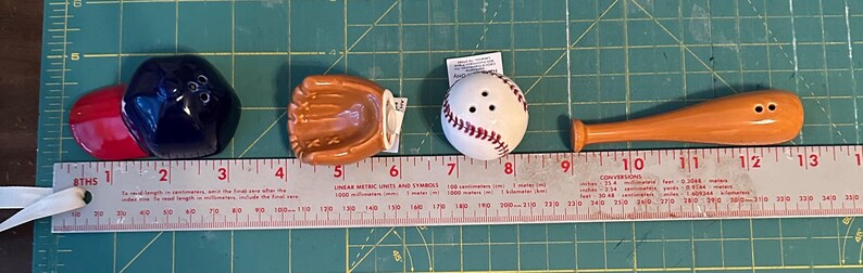 Puede incluir: Un juego de salero y pimentero con tem&aacute;tica de b&eacute;isbol. El juego incluye una gorra de b&eacute;isbol roja y azul marino, un guante de b&eacute;isbol marr&oacute;n, una pelota de b&eacute;isbol blanca con costuras rojas y un bate de b&eacute;isbol de madera.