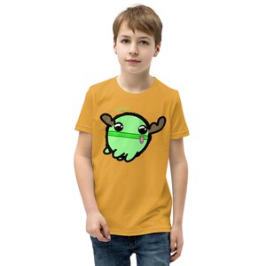 May include: Mustard yellow t-shirt featuring a cartoon green creature with brown antlers and a pink tongue. The creature has a green stripe across its face and a green halo. The shirt is worn by a person.