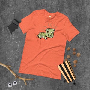 Op de afbeelding: Oranje T-shirt met een cartoon schildpad met een Frankenstein-achtige kop. De schildpad is groen en de kop is vierkant met steken en groene stoom. Het shirt heeft een effen oranje kleur.