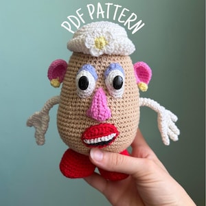 May include: A crocheted potato-shaped doll with a white hat, pink nose, red lips, and white teeth. It features purple eyebrows, pink ears, and white arms. The text PDF PATTERN is printed in white above the doll, which has red feet.