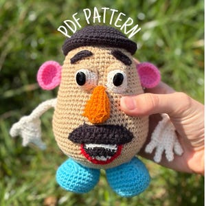 May include: A crocheted toy resembling a potato character with a tan body, black hat, pink ears, orange nose, black mustache, white and black eyes, red mouth, and blue feet. The text PDF PATTERN is printed in white above the toy's head.