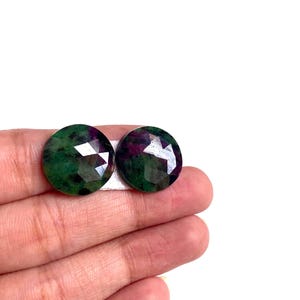 Puede incluir: Dos piedras preciosas redondas con superficies facetadas. Una es principalmente verde con inclusiones oscuras, la otra es una mezcla de verde y morado. Las gemas se sostienen sobre un fondo blanco.