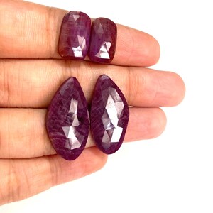May include: Four faceted, deep red gemstones with a polished finish. Two are rectangular, and two are teardrop-shaped. The gems are held in a hand against a white background, showcasing their color and cut.