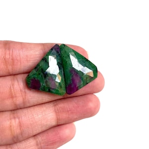 Könnte beinhalten: Zwei dreieckige Edelsteine mit facettierten Oberflächen. Die Edelsteine zeigen eine Mischung aus tiefgrünen und leuchtend roten Farbtönen mit einer polierten, reflektierenden Qualität. Die Steine werden in einer Hand vor weißem Hintergrund gehalten.