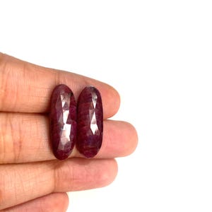 Può includere: Due pietre preziose di rubino sfaccettate a forma ovale. I rubini sono di colore rosso intenso con inclusioni interne visibili. Le pietre preziose sono tenute in mano su sfondo bianco. Questi rubini sono adatti per la creazione di gioielli.
