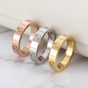 Könnte beinhalten: Drei Ringe in Roségold, Silber und Gold, jeweils mit einem sich wiederholenden kreuzförmigen Design. Die Ringe werden auf einer weißen Marmoroberfläche präsentiert, die ihre polierte Oberfläche und die verschiedenen Metalltöne hervorhebt.