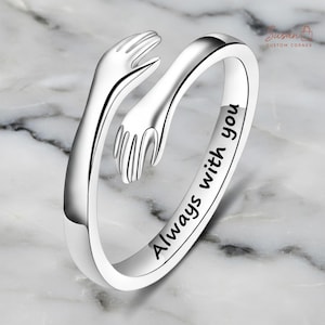 Könnte beinhalten: Ein silberner Ring mit zwei sich umarmenden Händen. Der Ring ist mit den Worten "Always with you" graviert. Der Ring liegt auf einer weiß-grauen Marmoroberfläche. Der Ring ist ein Schmuckstück.