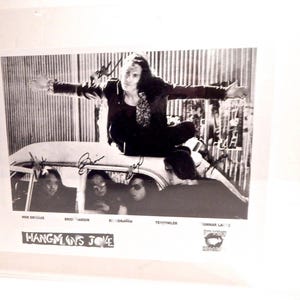 Puede incluir: Fotografía en blanco y negro de una banda posando sobre un coche. Los miembros de la banda están dentro y encima del coche. El nombre de la banda, "Hangman's Joke", está impreso en la parte inferior de la foto. La foto está firmada.