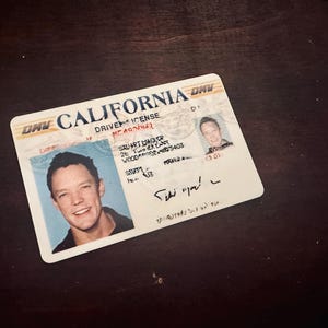 May include: A California driver's license with a photo of a smiling person. The license has the text "CALIFORNIA" and "DRIVER LICENSE" printed on it. The license is on a dark wooden surface.