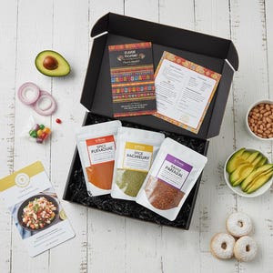 Puede incluir: Una caja negra llena de paquetes de especias, tarjetas de recetas e ingredientes como aguacate, frijoles y donas. Los paquetes de especias están etiquetados como "Spice Puemouae", "Spice Rachieuy" y "Rauri Papaual". La caja también contiene medio aguacate, cebolla roja en rodajas y caramelos.
