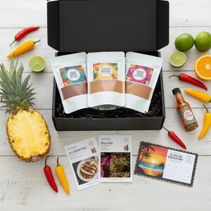 May include: A black box set containing three spice pouches, a bottle of hot sauce, and recipe cards. The set is surrounded by fresh ingredients like pineapple, limes, oranges, and chili peppers. The recipe cards feature images of tacos and other dishes.