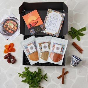 Puede incluir: Un juego de caja negra que contiene bolsas de especias, tarjetas de recetas y frutas secas. Las bolsas tienen diseños estampados coloridos. La tarjeta de recetas tiene el texto "Gusto Balsamico". El juego incluye palitos de canela, una pequeña taza azul y blanca y menta fresca.