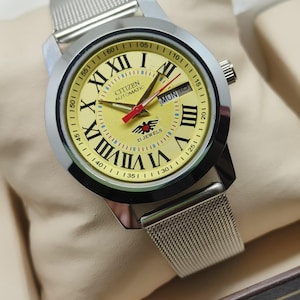 May include: A silver Citizen automatic wristwatch with a yellow face, black Roman numerals, and a mesh strap. The watch has red hands and a day indicator. The watch is displayed on a beige surface.