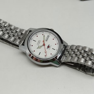 May include: A silver Citizen automatic wristwatch with a white face, black hour markers, and red accents. The watch has a stainless steel strap and shows the day of the week. A classic timepiece.
