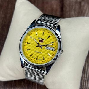 May include: A Seiko automatic wristwatch with a yellow dial and a silver-tone stainless steel mesh band. The watch face displays the day and date, with the word "SEIKO" at the top. The watch is on a white pillow.