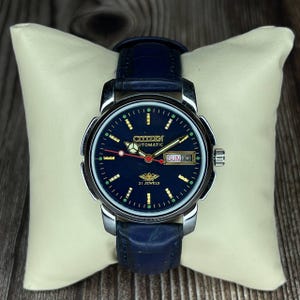 May include: A Citizen Automatic wristwatch with a dark blue face, gold hands, and a day-date display. The watch has a silver-coloured case and a dark blue leather strap. The watch is displayed on a cream-coloured cushion.