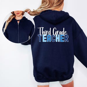 May include: Navy blue zip-up hoodie with the text "Third Grade TEACHER" in white and blue. The hoodie has a drawstring hood and a full-length zipper. The design is on the back of the hoodie.