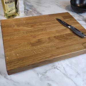May include: A rectangular wooden chopping board with a light brown finish. A stainless steel knife with a black handle rests on the board. The board is on a marble countertop, with a bottle of olive oil and a black bowl in the background.