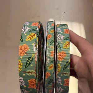 May include: Several spools of green ribbon featuring a repeating pattern of orange, yellow, and white autumn leaves. Ideal for crafts, gift wrapping, or seasonal decor. The detailed leaf design adds a touch of autumnal charm to any project.