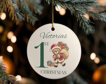 May include: A round, off-white Christmas ornament with the text "Victorias 1st Christmas" in green script. A cartoon teddy bear in a Santa suit carries a sack of gifts. The ornament hangs from a gold string.