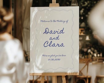 May include: A wooden easel displays a sign with the words "Welcome to the Wedding of David and Clara." The sign is white with blue script and a decorative border. The date 01.30.2030 is also visible. The scene is outdoors, with a blurred background.