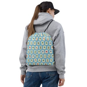 May include: A light blue drawstring bag with a pattern of Mahjong tiles. The tiles are white with red, green, and black symbols and characters. The bag has black drawstrings and is worn over the shoulders.