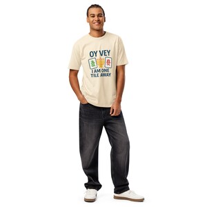 May include: A person wearing a cream-colored t-shirt with the text "OY VEY I AM ONE TILE AWAY" in navy blue and teal. The shirt features a graphic of Mahjong tiles and a menorah. The person is wearing black jeans and white sneakers.