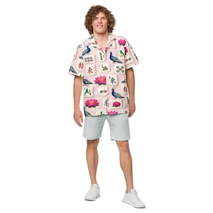 May include: A short-sleeved button-down shirt with a pink background, featuring a pattern of Mahjong tiles, peacocks, and lotus flowers. The person is also wearing light blue shorts and white sneakers.