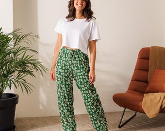May include: A person wearing a white crop top and green pajama pants. The pajama pants have a repeating pattern of playing cards in white, red, and black on a green background. The person is standing in front of a brown chair.