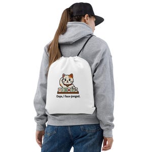 May include: White drawstring bag with a cartoon cat design, looking forlorn, and mahjong tiles. The text "Oops, I faux-jonged" is printed below the cat. The bag has black drawstrings. A fun accessory.