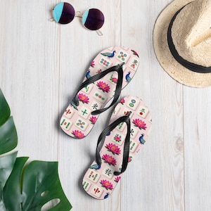 May include: Pink flip-flops with a Mahjong tile pattern, featuring lotus flowers, peacocks, and Chinese characters. The flip-flops have black straps. Sunglasses and a straw hat are also visible.