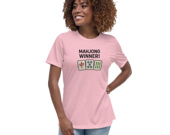 May include: A pale pink t-shirt featuring the words "MAHJONG WINNER!" and three illustrated Mahjong tiles in green, white, and red. The shirt has a relaxed fit.
