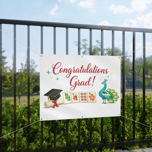 Könnte beinhalten: Weißes Banner mit dem Text "Congratulations Grad!" in roter Schrift. Das Banner zeigt eine Abschlusskappe, ein Diplom und eine Pfau-Illustration. Das Banner hängt an einem schwarzen Metallzaun.