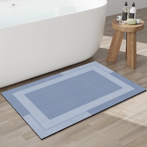 May include: A rectangular blue bath mat with a light blue border and a darker blue center. The mat is placed on a wooden floor near a white bathtub and a small wooden stool with toiletries.