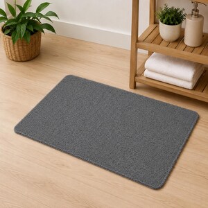 May include: A rectangular dark gray bath mat with a textured, plush surface and rounded corners. The mat features a dense, soft pile construction and is positioned flat on a light-colored wooden floor, showcasing a simple and functional design.