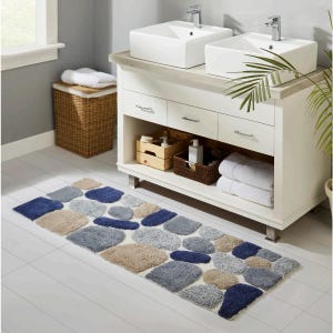 May include: A bathroom rug with a stone design in shades of blue, gray, and beige. The rug is rectangular and placed on a white tiled floor in front of a white vanity with two sinks. A wicker hamper and towels are also visible.