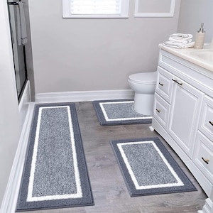 May include: A three-piece bathroom rug set. The rugs are gray with a white border. The set includes a long rectangular rug, a square rug, and a smaller rectangular rug. The rugs are placed on a gray wooden floor.