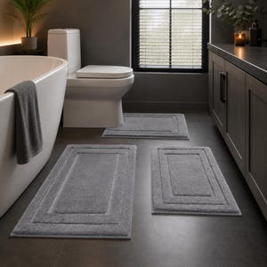 May include: A set of three rectangular gray bath mats with a textured border design. The mats are arranged on a dark floor in a bathroom setting, featuring a plush, soft surface texture and a consistent solid gray color throughout each piece.