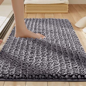 May include: A gray bath mat with a textured, bubble-like surface. The rectangular mat is on a wooden floor, with a person's foot stepping onto it. A wicker basket and slippers are in the background.
