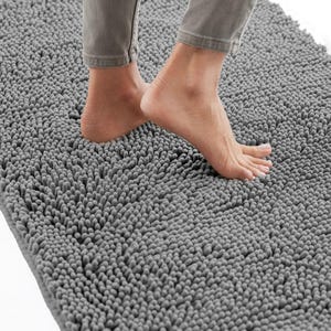 May include: A gray bath mat with a textured, shaggy surface. The mat is rectangular and appears soft. A person's feet are visible, standing on the mat. The mat is designed for use in a bathroom.
